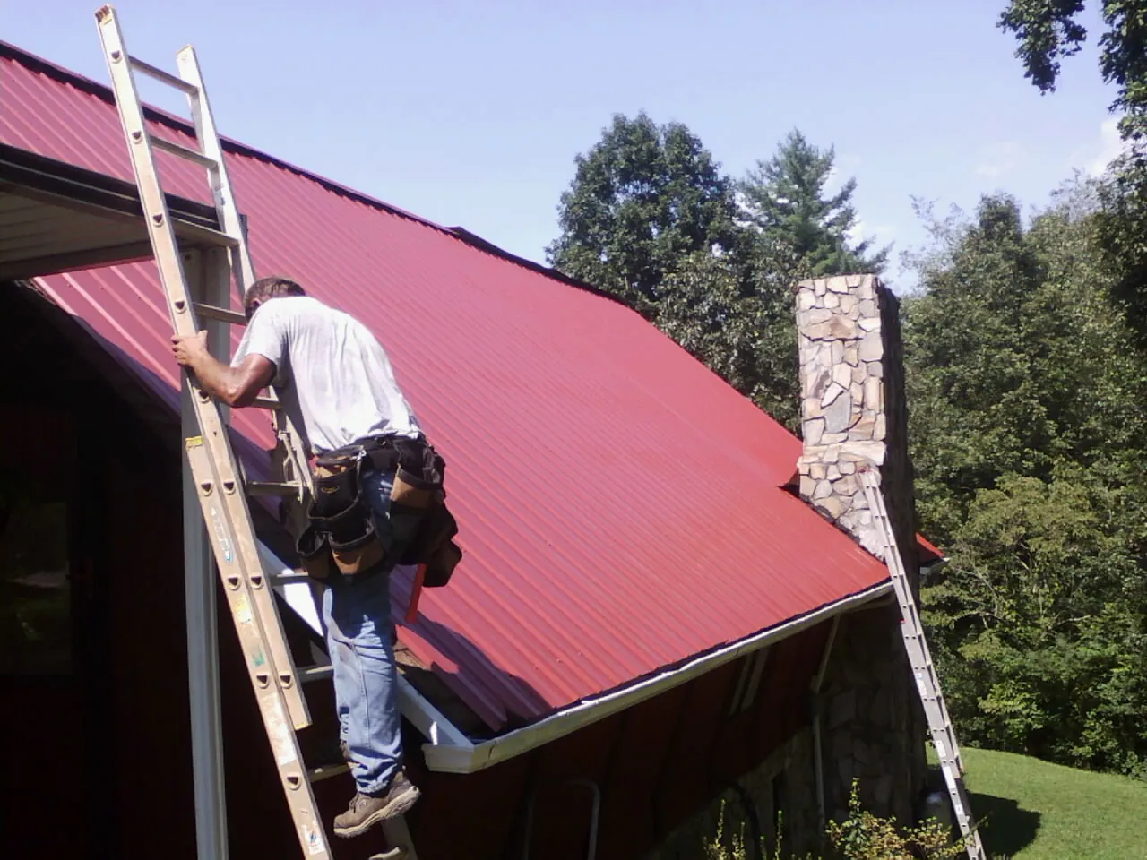 Roofer on a ladder beside a red metal roof and stone chimney — Soffit & Fascia Repair by Roofing Brothers in Clatskanie